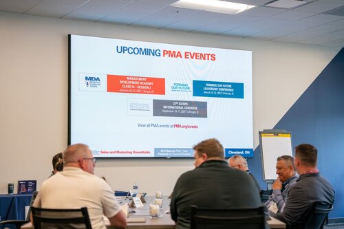 The image depicts a professional meeting or conference setting with a group of individuals seated around a table. The room is well-lit with a modern design, featuring a large screen displaying information about upcoming events. The screen prominently shows the title "UPCOMING PMA EVENTS" in bold letters, with a focus on events related to management and leadership development, such as the "MANAGEMENT DEVELOPMENT ACADEMY" and "FORMING OUR FUTURE LEADERSHIP CONFERENCE." The screen also mentions the "21st ICOSPA INTERNATIONAL CONGRESS."

Participants at the table are engaged in discussion, suggesting a collaborative atmosphere. The individuals are dressed in business casual attire, indicating a professional yet relaxed environment. On the table, there are nameplates, notepads, and water bottles, typical of a conference or meeting setup.

In the background, a flip chart is visible, further emphasizing the business context. The room's decor includes a blue and white color scheme, contributing to a clean and corporate ambiance. The setting is likely in Cleveland, OH, as indicated by the text on the screen.

Overall, the image conveys a sense of professional development and networking, with a focus on future-oriented leadership and management training events.