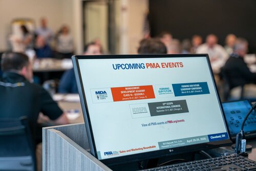 The image depicts a professional conference or meeting setting. In the foreground, a computer monitor displays a slide titled "UPCOMING PMA EVENTS" with a list of events. The events include:

1. "MANAGEMENT DEVELOPMENT ACADEMY CLASS 57 SESSION 1" scheduled for January 26-27, 2023.
2. "FORMING OUR FUTURE LEADERSHIP CONFERENCE" set for March 15-17, 2023.
3. "ICOSPA INTERNATIONAL CONGRESS" on September 24-27, 2023.

The bottom of the slide mentions "Visit pma.org/events #PMAjourneys" and provides location details: "Cleveland, OH" and "Sales and Marketing Roundtable" with dates "April 28-29, 2020."

In the background, there are several people seated at tables, suggesting a workshop or seminar environment. The setting appears to be a conference room with a focus on business or professional development. The atmosphere is formal and focused, with participants engaged in discussions or listening attentively.

The lighting is bright and conducive to a professional setting, with neutral tones dominating the room. The image conveys a sense of organization and professional growth, emphasizing networking and learning opportunities.