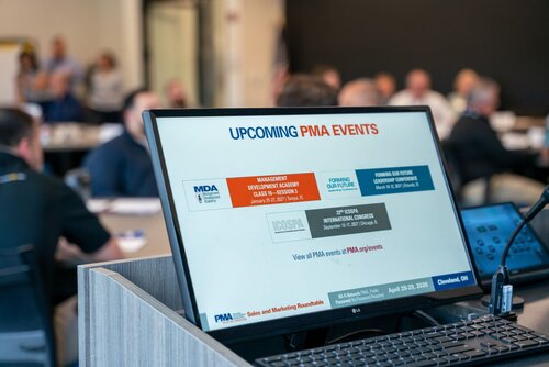 The image depicts a professional conference or meeting setting, likely a business or industry event. In the foreground, there is a computer monitor displaying a presentation slide. The slide prominently features the title "UPCOMING PMA EVENTS" and includes details about various events such as the "Management Development Academy" and "Forming Our Future Leadership Conference." The slide also contains logos, including "MDA" and "ICOSPA," and a web address for more information.

The setting appears to be a conference room with several people seated at tables in the background, engaged in discussions or listening to a presentation. The room is well-lit, creating a professional and focused atmosphere. The predominant colors on the screen are blue, red, and white, which are typical for corporate presentations.

The overall theme suggests a formal business environment, with an emphasis on industry-specific events and networking opportunities. The image conveys a sense of organization and professionalism, typical of corporate gatherings or industry conferences.
