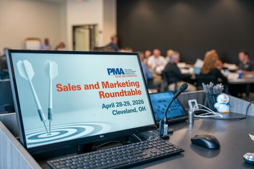 The image depicts a professional setting, likely a conference or business meeting. In the foreground, there is a computer monitor displaying a slide with the text "Sales and Marketing Roundtable," scheduled for "April 28-29, 2026, Cleveland, OH." The slide includes the logo of the "PMA Precision Marketing Association" and features a graphic of two darts hitting a target, symbolizing precision and focus.

The setting appears to be a conference room with a modern, professional atmosphere. On the desk near the monitor, there is a microphone, suggesting that presentations or discussions are taking place. Other items on the desk include a pen holder with pens and a small figurine, adding a personal touch to the workspace.

In the background, several people are seated at tables, engaged in conversation or listening attentively, indicating an interactive and collaborative environment. The room has a neutral color palette with predominantly gray and black tones, contributing to a formal and business-like ambiance.

The overall theme of the image conveys a sense of professionalism, collaboration, and focus on marketing and sales strategies. The presence of the PMA logo and the event details suggest an organized and structured gathering aimed at industry professionals.