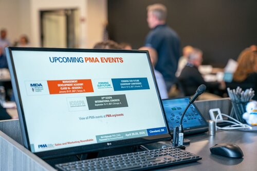 The image depicts a professional setting, likely a conference or seminar, focused on upcoming events related to PMA (Precision Metalforming Association). The foreground features a computer monitor displaying a slide titled "UPCOMING PMA EVENTS" with details of several events:

1. **Management Development Academy Class 57 - Session 1** scheduled for January 25-27, 2023, in Tampa, FL.
2. **Forming Our Future Leadership Conference** on March 5-8, 2023, in New Orleans, LA.
3. **ICOSPA International Congress** from September 17-20, 2023, in Cologne, Germany.

The slide includes a call to action to view all PMA events at a specified URL, with emphasis on "PMA.org/events."

In the background, there are blurred figures, suggesting a gathering of people, possibly attendees or participants in a meeting or presentation. The setting appears to be a conference room, with a focus on business or industry-specific discussions.

The predominant colors on the screen are blue, orange, and gray, which are often associated with professionalism and corporate branding. The lighting is bright, typical of indoor conference environments, enhancing visibility and focus on the presentation.

The atmosphere conveys a sense of organization and professionalism, with an emphasis on networking and industry advancement. The presence of a microphone and control panel on the desk suggests that this is a speaker's podium or a presentation area, reinforcing the formal and informative nature of the event.

Overall, the image captures a moment in a business conference or seminar, highlighting upcoming industry events and the collaborative environment typical of such gatherings.
