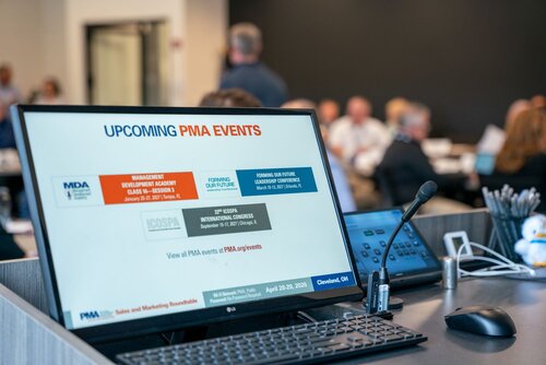 The image depicts a professional setting, likely a conference or seminar, with a focus on a computer monitor displaying information about upcoming events. The screen prominently features the text "UPCOMING PMA EVENTS" in bold, with a list of events beneath it. These events include:

1. "MANAGEMENT DEVELOPMENT ACADEMY CLASS 75 - SESSION 1" scheduled for January 25-27, 2023.
2. "FORMING OUR FUTURE LEADERS CONFERENCE" scheduled for March 7-8, 2023.
3. "STAMPING INNOVATION TOUR" scheduled for April 12-13, 2023.

The bottom of the screen includes a URL: "View all PMA events at PMA.org/events."

The setting is a modern conference room with several people seated at tables in the background, engaged in what appears to be a meeting or discussion. The room is well-lit, with a neutral color palette dominated by whites and grays, contributing to a professional and focused atmosphere.

In the foreground, there is a microphone and a pen holder with various writing instruments, suggesting a speaker's podium or presentation area. The overall ambiance is one of professionalism and organization, suitable for a business or industry-related event.

The image captures the essence of a corporate environment, emphasizing planning and information dissemination, typical of business conferences or industry seminars.