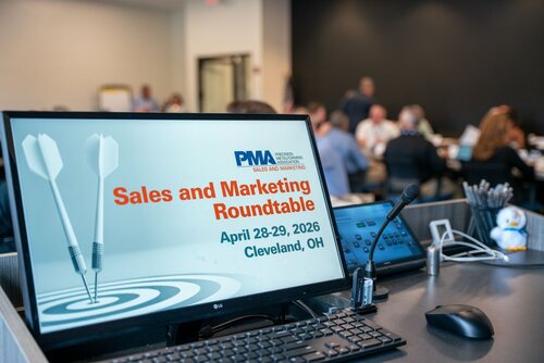 The image depicts a professional setting, likely a conference or seminar, focused on sales and marketing. In the foreground, a computer monitor displays a slide with the text "Sales and Marketing Roundtable," scheduled for "April 28-29, 2026, Cleveland, OH." The slide includes the logo of an organization, "PMA," which stands for "Precision Metalforming Association," indicating the industry focus of the event. The slide also features a graphic of two darts hitting a target, symbolizing precision and goal achievement.

The setting appears to be a conference room with a modern, professional atmosphere. On the desk in front of the monitor, there is a microphone, suggesting that presentations or discussions are taking place. Additional items include a phone and a small penguin figurine, adding a touch of personality to the workspace.

In the background, several people are gathered around tables, engaged in conversation or listening attentively, indicating an interactive or collaborative environment. The lighting is bright and conducive to a professional setting, with a mix of natural and artificial light. The overall atmosphere is one of focused engagement, typical of a business meeting or industry roundtable.

The image conveys a sense of professionalism and industry-specific networking, emphasizing themes of collaboration, strategic planning, and goal-oriented discussions within the sales and marketing sectors.