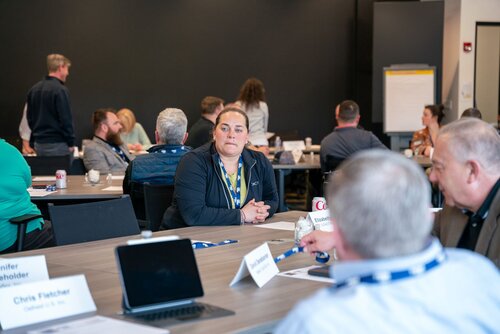 The image depicts a professional meeting or conference setting, with several individuals seated around tables in a well-lit room. The atmosphere is collaborative and focused, suggesting a workshop or seminar environment. 

Prominent elements include:

1. **Participants**: Individuals are engaged in discussion, with some seated and others standing. They appear to be actively listening or conversing, indicating a group activity or breakout session.

2. **Name Tags and Lanyards**: Many participants wear lanyards with name tags, suggesting an organized event where identification is necessary. Some name tags are visible, with names like "Jennifer Stakeholder" and "Chris Fletcher."

3. **Technology**: A laptop is open on one of the tables, indicating the use of technology for note-taking or presentations.

4. **Tables and Materials**: The tables are equipped with papers, pens, and drinks, such as a can of Coca-Cola, implying a long-duration event where refreshments are provided.

5. **Presentation Tools**: In the background, there is a flip chart, which is commonly used for presentations or brainstorming sessions.

6. **Color and Lighting**: The room is brightly lit, with neutral tones dominating the scene, contributing to a professional and formal atmosphere.

The setting suggests a corporate or educational context, possibly a training session, business meeting, or conference. The presence of name tags and organized seating implies a structured event with multiple participants engaging in professional development or collaborative activities.