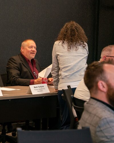 The image depicts a professional setting, likely a meeting or conference. A man is seated at a table, engaged in conversation with a standing woman. The man is wearing a dark suit and a red shirt, and he appears to be smiling, suggesting a friendly or positive interaction. In front of him is a nameplate that reads "Mark Fortuna, President, Association," indicating his role and possibly the context of the event as a formal meeting or association gathering. A can of Coca-Cola is placed on the table near him, adding a casual element to the setting.

The woman is dressed in a casual outfit, including a light-colored sweater, and is facing away from the camera, engaging with the seated man. The background shows a dark wall, contributing to a professional and subdued atmosphere. Other individuals are partially visible, seated around the table, suggesting a group discussion or presentation.

The lighting is bright, focusing on the individuals and the table, creating a clear and focused scene. The overall atmosphere is professional yet approachable, indicative of a formal event with a relaxed tone. The setting suggests a conference room or similar environment, where business or organizational matters are being discussed.