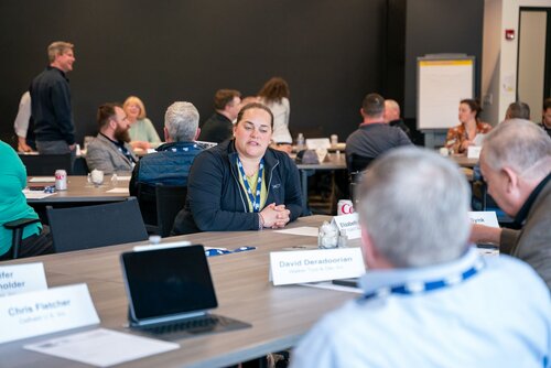 The image depicts a professional meeting or workshop setting, likely taking place in a conference room. Several individuals are seated around tables, engaged in conversation. The atmosphere is collaborative and focused, suggesting a discussion or brainstorming session. 

Prominent objects include laptops, notepads, and nameplates, indicating a formal meeting environment. The nameplates on the tables display names such as "David Deradoorian" and "Chris Fletcher," adding a personalized touch to the setting. A whiteboard or flip chart is visible in the background, often used for presentations or notes during meetings.

The room is well-lit with natural and artificial lighting, creating a bright and inviting atmosphere. The participants are casually dressed, suggesting a relaxed yet professional environment. The predominant colors in the image are neutral tones, with the black walls and furniture contrasting with the lighter colors of the participants' clothing.

The image conveys a sense of teamwork and communication, common in corporate or educational settings. It captures the essence of a modern professional gathering, where ideas are exchanged, and collaboration is key.