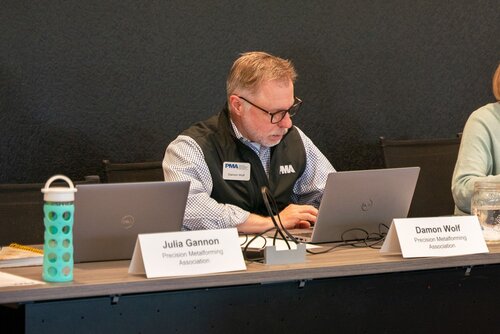 The image depicts a professional setting, likely a conference or meeting, featuring a man seated at a table. He is focused on his laptop, suggesting engagement in work or a presentation. The man is wearing glasses and a black vest with a name tag that reads "Damon Wolf" and "PMA," indicating his association with the Precision Metalforming Association. 

To his left, there is another laptop and a nameplate for "Julia Gannon," also from the Precision Metalforming Association. The table is equipped with typical meeting items, including a turquoise water bottle with a polka dot pattern and a glass of water, indicating a long session or presentation.

The background is a simple, dark wall, which keeps the focus on the individuals and their activities. The lighting is bright and even, creating a professional and focused atmosphere. The setting suggests a formal meeting or workshop related to the metalforming industry, emphasizing collaboration and information sharing.