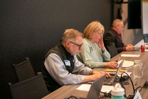 The image depicts a professional setting, likely a meeting or workshop, with three individuals seated at a long table. The environment suggests a conference room, characterized by a neutral, corporate atmosphere with dark walls and simple decor. 

The first individual, a man with glasses, is focused on a laptop. He wears a name tag and a black vest over a checkered shirt, suggesting a business-casual dress code. The second individual, a woman, is engaged with a phone, possibly checking messages or emails. She wears a light blue sweater and a pearl necklace, indicating a professional appearance. The third person, another man, is partially visible and appears to be reading or reviewing documents.

On the table, there are various items including laptops, notebooks, a water bottle, and a can of soda, indicating a work session or meeting. The lighting is bright and even, providing a clear view of the participants and their activities.

The overall atmosphere is one of concentration and productivity, typical of a business meeting or workshop. The setting and actions suggest a focus on collaboration and communication, with technology playing a central role in the activities depicted.