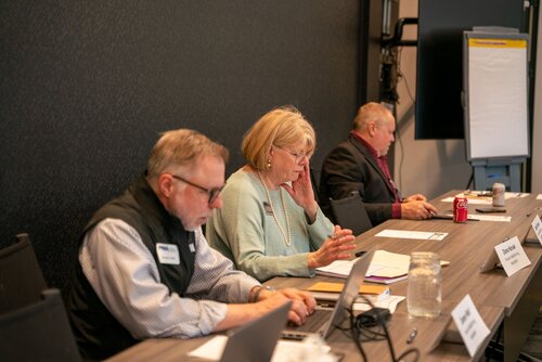 The image depicts a professional meeting or conference setting. Three individuals are seated at a long table, each engaged in their tasks. The setting appears to be a conference room with a dark, textured wall as the backdrop. 

On the left, a man with glasses is focused on his laptop, wearing a vest over a shirt. In the center, a woman with blonde hair is wearing a light blue sweater and is engaged in a phone call, with her hand resting on her cheek. She has a notepad and pen in front of her, indicating active participation in the meeting. On the right, another man is taking notes with a pen, and a can of Diet Coke is visible beside him, suggesting a casual aspect to the professional environment.

The table is equipped with nameplates, suggesting a formal meeting or panel discussion. A flip chart stand is visible in the background, indicating potential presentations or brainstorming sessions. The lighting is bright and even, contributing to a focused and business-like atmosphere.

The overall mood is one of concentration and professionalism, with each individual engaged in their respective tasks. The presence of technology, such as laptops and phones, highlights a modern, connected work environment.