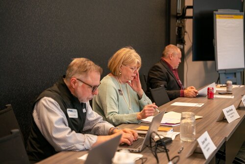 The image depicts a professional meeting or conference setting with three individuals seated at a long table. The individuals appear to be engaged in a focused discussion or taking notes. 

- **Individuals**: 
  - The person on the left is wearing glasses and a vest with a visible name tag, suggesting a formal or business environment.
  - The middle individual, a woman, is wearing a light-colored sweater and is writing in a notebook, indicating active participation.
  - The person on the right is dressed in a suit and is looking down, possibly reviewing documents or notes.

- **Objects and Setting**:
  - Laptops and notebooks are present, indicating a work-related context.
  - A can of Coca-Cola is on the table, adding a casual element to the otherwise formal setting.
  - A flip chart is visible in the background, suggesting a presentation or brainstorming session.
  - The room has a modern, professional appearance with a dark wall and a large screen or monitor.

- **Atmosphere**:
  - The lighting is bright and even, creating a focused and professional atmosphere.
  - The participants appear concentrated and engaged, conveying a serious and business-like mood.

- **Cultural/Contextual Elements**:
  - The setting and attire suggest a corporate meeting or workshop, possibly related to business strategy or planning.

This image captures a typical scene from a business meeting, emphasizing collaboration and professional engagement.