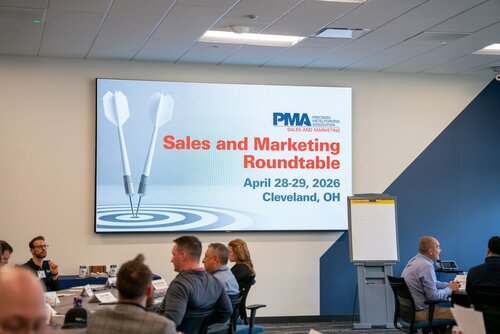 The image depicts a professional setting, likely a conference or business meeting. The focal point is a large screen displaying the text "Sales and Marketing Roundtable" along with the dates "April 28-29, 2026" and the location "Cleveland, OH." The screen also features the logo "PMA," which stands for Precision Metalforming Association, and an image of two darts hitting a target, symbolizing precision and goal achievement.

In the foreground, several individuals are seated around tables, engaged in discussions or listening attentively. The tables are equipped with nameplates, papers, and water bottles, indicating a formal meeting environment. The attendees are dressed in business attire, contributing to the professional atmosphere.

The room is well-lit with a modern design, featuring a ceiling with recessed lighting and a neutral color palette dominated by blues and grays. A flip chart stand is visible to the right, suggesting interactive sessions or presentations may be part of the event.

The overall atmosphere is focused and collaborative, emphasizing themes of business strategy and professional development. The setting suggests a gathering of industry professionals aimed at networking and sharing insights in sales and marketing.
