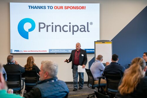 The image depicts a professional setting, likely a conference or seminar, focused on sales and marketing. At the center, a speaker stands in front of an audience, holding a piece of paper, and appears to be delivering a presentation. The audience is seated at tables, attentively listening.

Prominently displayed behind the speaker is a large screen featuring the text "THANKS TO OUR SPONSOR!" in blue and red, followed by the logo and name "Principal" in gray with a blue circular symbol. This suggests that Principal is a key sponsor of the event. The screen also includes logos for "PMA" and "Cleveland," indicating possible affiliations or locations related to the event.

The setting is a modern conference room with a neutral color palette, featuring blue and gray tones. The lighting is bright and professional, enhancing the clarity of the presentation. There is a flip chart to the right of the speaker, suggesting interactive or collaborative elements in the session.

The atmosphere is formal and educational, with a focus on business networking and knowledge sharing. The presence of multiple individuals in business attire indicates a professional and corporate environment.