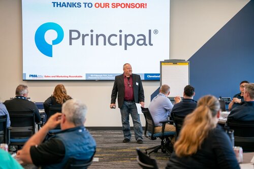 The image depicts a professional setting, likely a business conference or seminar. At the center, a man stands confidently in front of a large screen displaying the logo and name "Principal" with a stylized blue and teal "P" symbol. Above the logo, red text reads, "THANKS TO OUR SPONSOR!" indicating sponsorship recognition.

The setting is a modern conference room with a neutral color palette, featuring gray and blue tones. The room is filled with attendees seated at tables, suggesting a roundtable or workshop format. They are casually dressed, indicative of a business-casual environment, and are engaged in the presentation.

The speaker, dressed in a blazer and jeans, holds a paper, suggesting he is either presenting or facilitating the session. A flip chart is positioned to the right of the screen, ready for use, enhancing the interactive nature of the event.

The atmosphere is professional and focused, with an emphasis on learning and collaboration. The presence of branding and sponsorship highlights the business-oriented theme of the event.