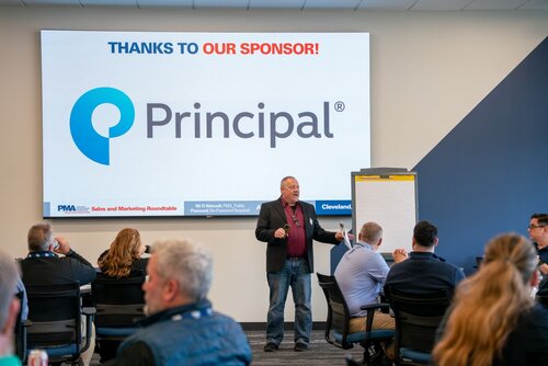The image depicts a professional event or conference setting, likely a sales and marketing roundtable, as indicated by the text on the screen. The main focus is a man standing in front of a large screen, engaging with an audience seated at tables. The screen prominently displays the logo and name "Principal," acknowledging them as a sponsor with the text "THANKS TO OUR SPONSOR!" in red and blue. 

The setting is a modern conference room with neutral-colored walls and carpeted flooring. Attendees are dressed in business casual attire, suggesting a formal yet relaxed atmosphere. The audience appears attentive, with some individuals taking notes on a flip chart positioned to the right of the speaker. 

The lighting is bright and even, typical of indoor conference environments, enhancing visibility and focus on the speaker and screen. The overall atmosphere is professional and collaborative, indicative of a networking or educational event.