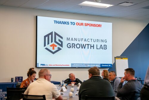 The image depicts a professional setting, likely a conference or business meeting, focused on manufacturing. The central feature is a large screen displaying the text "THANKS TO OUR SPONSOR!" in bold letters, followed by the logo and name "Manufacturing Growth Lab." The logo consists of stylized letters, incorporating an industrial or technical design element.

Below the main text, a banner displays details about the event: "PMA Sales and Marketing Roundtable," with the location "Cleveland, OH" and dates "April 28-29, 2023." This suggests a formal gathering of industry professionals, possibly for networking or educational purposes.

In the foreground, several individuals are seated around a conference table, engaged in discussion. The table is equipped with nameplates, papers, and small water bottles, indicating an organized and structured meeting. The participants appear focused and attentive, contributing to a serious and professional atmosphere.

The room is well-lit, with a modern design featuring neutral colors like white and blue, contributing to a corporate and efficient ambiance. A flip chart stands near the screen, hinting at presentations or collaborative activities.

Overall, the image conveys a sense of industry collaboration and professional development within the manufacturing sector.