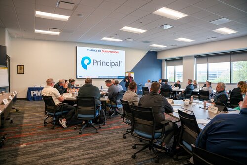 The image depicts a professional meeting or conference setting within a modern office environment. Attendees are seated around a large, U-shaped arrangement of tables, facing a presentation screen at the front of the room. The screen displays a slide with the text "THANKS TO OUR SPONSOR!" followed by the logo and name "Principal," suggesting a sponsorship acknowledgment.

The room is well-lit with a combination of natural light streaming through large windows on the right and overhead fluorescent lighting. The walls are painted in neutral tones, contributing to a professional atmosphere. Attendees are dressed in business casual attire, indicating a formal yet relaxed meeting environment.

On the left side of the room, a table with a blue tablecloth displays the acronym "PMI," which could reference the Project Management Institute, suggesting a connection to project management or related professional development activities.

The carpet features a modern design with a mix of gray and red tones, adding a contemporary touch to the room's decor. The overall atmosphere is focused and collaborative, with participants appearing engaged in the presentation or discussion.

This setting is typical for corporate training sessions, workshops, or seminars, where networking and knowledge sharing are key components. The presence of the sponsor's logo and the professional setup indicate an organized and possibly recurring event.