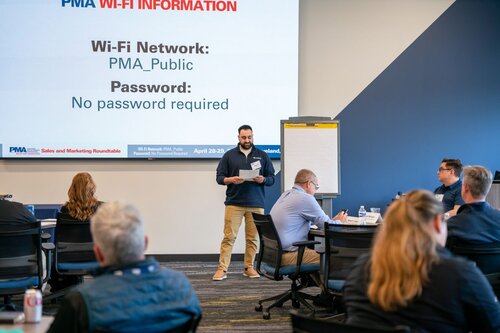 The image depicts a professional setting, likely a conference or workshop, focused on a presentation. The room is filled with individuals seated at tables, suggesting a collaborative or educational event. A man stands at the front, holding papers, and appears to be speaking or presenting to the audience. 

The background features a large screen displaying text related to Wi-Fi information, specifically:

- "Wi-Fi Network: PMA_Public"
- "No password required"

This indicates the event's location provides open internet access for attendees. The screen also includes logos and text related to the event, such as "PMA Sales and Marketing Roundtable" and dates "April 28 & 29."

The room's decor includes a modern, professional aesthetic with a blue and gray color scheme. Attendees are dressed in business casual attire, enhancing the professional atmosphere. A flip chart is visible beside the presenter, indicating potential use for notes or additional information.

The setting conveys a formal yet approachable environment, emphasizing learning, networking, and information sharing. The presence of a soda can on one of the tables suggests a casual aspect to the event, allowing for refreshments during sessions. Overall, the image captures a moment of engagement and communication within a business context.