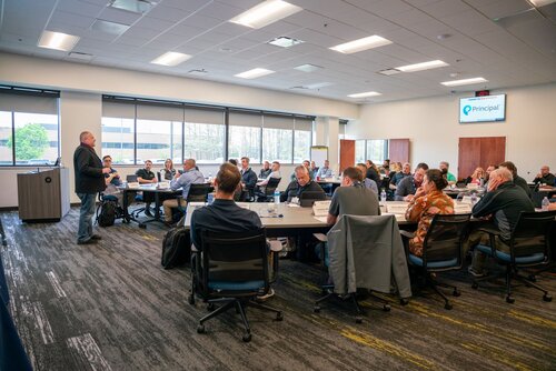 The image depicts a professional meeting or conference taking place in a modern, well-lit conference room. The room is spacious, with large windows allowing natural light to illuminate the space. Approximately twenty individuals are seated around several rectangular tables arranged in a U-shape, facilitating interaction and discussion. 

A speaker stands at the front of the room near a podium, addressing the attendees. The audience appears attentive, with some individuals taking notes or looking towards the speaker. The attire of the participants is business casual, indicating a formal yet relaxed atmosphere.

On the wall behind the speaker, there is a large screen displaying the logo and name "Principal," suggesting the event might be organized by or for this company. The room features modern office furniture, including ergonomic chairs and tables with a light wood finish. 

The setting suggests a corporate training session, seminar, or workshop. The atmosphere is professional and focused, with a sense of collaboration and learning. The presence of technology, such as laptops and a projector, indicates a contemporary business environment.