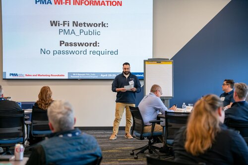 The image depicts a professional meeting or conference setting. A man stands at the front of a room, holding a piece of paper, and appears to be speaking to an audience seated at tables. The room is arranged with several round tables, each accommodating a few attendees who are listening attentively. The atmosphere is formal and focused, indicative of a business or educational seminar.

Prominently displayed on a screen behind the speaker is text providing Wi-Fi information: "Wi-Fi Network: PMA_Public" and "Password: No password required." This suggests the event is organized by or associated with an entity abbreviated as PMA. Additional branding on the screen includes "PMA Sales and Marketing Roundtable" and a date range, "April 28-29," indicating a multi-day event.

The setting is well-lit with natural and artificial lighting, contributing to a professional and welcoming environment. The color scheme includes neutral tones with accents of blue and white, enhancing the formal atmosphere. Attendees are dressed in business casual attire, further emphasizing the professional context.

A flip chart is positioned near the speaker, suggesting the potential for interactive discussions or presentations. The overall mood is one of engagement and information exchange, typical of a corporate or industry-specific gathering.