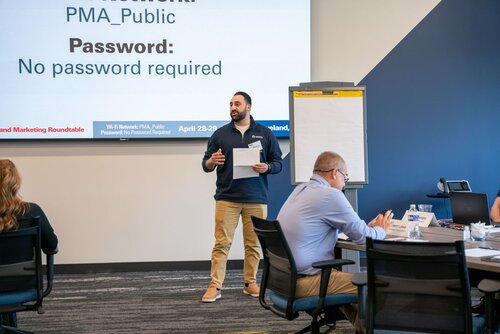 The image depicts a professional setting, likely a conference or workshop. A man stands at the front of a room, holding a piece of paper, possibly delivering a presentation or leading a discussion. He is dressed in a casual yet professional manner, wearing a dark sweater and khaki pants. 

Behind him, a large screen displays text with details about a Wi-Fi network: "Network: PMA_Public" and "Password: No password required." This suggests the event is organized and provides internet access for attendees. The screen also mentions "April 28-29," indicating the event's dates.

To the right of the speaker, there is a flip chart, which may be used for notes or brainstorming during the session. The room has a modern, corporate design with a blue and gray color scheme, and the carpet features a patterned design.

Several attendees are seated at tables, engaged and possibly taking notes. The tables are equipped with water bottles and notepads, emphasizing a focus on comfort and productivity. The atmosphere is professional and collaborative, typical of a business or educational seminar.

Overall, the image conveys a sense of organized, interactive learning or discussion, likely aimed at professionals or industry participants.