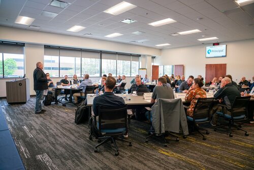 The image depicts a professional meeting or conference taking place in a modern, well-lit conference room. The room is filled with individuals seated around large, rectangular tables arranged in a U-shape, allowing for interaction and discussion. A presenter stands at the front, addressing the group, suggesting a presentation or lecture format. 

The room features large windows on one side, allowing natural light to illuminate the space, complementing the overhead fluorescent lighting. The walls are painted in neutral tones, contributing to a professional atmosphere. The floor is carpeted with a pattern that combines dark and light shades, adding a touch of sophistication.

Prominent in the image is a digital screen displaying the logo and name "Principal," indicating either the name of the event, the organization hosting it, or the company involved. Participants are dressed in business casual attire, reflecting a formal yet relaxed business setting. Some attendees have laptops and notepads, suggesting active engagement and note-taking.

The overall atmosphere is one of focused professionalism, with participants appearing attentive and engaged in the proceedings. The setting suggests a corporate training session, seminar, or business meeting, characterized by collaboration and information exchange.