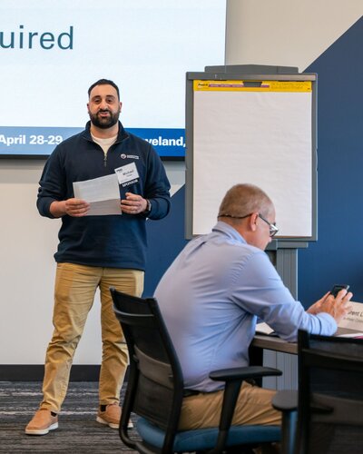 The image depicts a professional setting, likely a conference or workshop. In the foreground, a man is standing and holding papers, appearing to be presenting or speaking to an audience. He is dressed in a casual yet professional manner, wearing a navy blue pullover and khaki pants. Behind him is a large flip chart on a stand, which is blank, suggesting a session in progress or preparation for note-taking.

Another individual is seated at a table, focused on a smartphone, indicating a possible break or multitasking during the event. This person is wearing a light blue shirt and glasses, contributing to the professional atmosphere.

The background features a large screen with partial text visible, hinting at a presentation or agenda. The text includes a date, "April 28-29," and a location, "Cleveland," suggesting the timing and venue of the event. The setting is modern, with a neutral color palette of blues and grays, creating a calm and focused environment.

The overall atmosphere is one of learning and collaboration, typical of business or educational gatherings. The presence of technology and presentation tools emphasizes a structured and informative context.