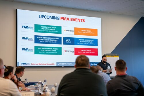 The image depicts a professional conference or seminar setting. At the forefront, a man is standing near a flip chart, seemingly presenting or discussing with attendees seated around a table. The attendees are engaged, taking notes or listening attentively.

In the background, a large screen displays information about upcoming events organized by PMA (Precision Metalforming Association). The screen lists several events with dates and locations, such as:

- "Press Capacity, Set Up and Safety Seminar" on May 9, 2023, in Cleveland, OH.
- "Human Resources and Training Professionals Roundtable" on May 11, 2023, in Cleveland, OH.
- "OSHA for Mechanical Presses & PFA/F Webinar Series" on May 23, 2023, in Webinar Series.
- "FABTECH Mexico" from May 16-18, 2023, in Mexico City.
- "One Voice in Washington: Public Policy Update" on May 24, 2023, in Webinar Series.
- "FABTECH Canada" from June 14-16, 2023, in Toronto.

The setting is a modern conference room with a neutral color palette dominated by blues and whites, contributing to a professional and focused atmosphere. The lighting is bright and conducive to a learning environment.

The image conveys a sense of organized professional development and networking, typical of industry-specific conferences. The presence of PMA and FABTECH suggests a focus on manufacturing and metalforming industries. The event is likely aimed at professionals seeking to enhance their knowledge and skills in these fields.