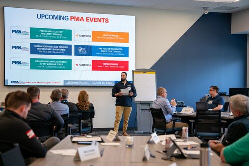 The image depicts a professional meeting or conference setting with a focus on upcoming events organized by PMA (Precision Metalforming Association). The scene is set in a modern conference room with several attendees seated around a large table, listening to a speaker standing at the front. The speaker is holding a microphone and appears to be presenting information from a large screen behind him.

The screen displays a slide titled "UPCOMING PMA EVENTS" with a list of events, including:

1. "Press Capacity Set Up and Safety Seminar" in Cleveland, OH.
2. "Human Resources and Training Professionals Roundtable" in Cleveland, OH.
3. "OSHA for Mechanical Presses" in Cleveland, OH.
4. "FABTECH Mexico" in Monterrey, Mexico.
5. "Die Maintenance Process Seminar" in Cleveland, OH.
6. "FABTECH Canada" in Toronto, Canada.

Each event listing includes dates and locations, and some feature logos of associated organizations, such as FABTECH and One Voice.

The room is well-lit with natural and artificial lighting, creating a professional and focused atmosphere. Attendees are engaged, with some taking notes or looking at the presentation. The setting suggests a business or industry-related event, emphasizing networking and knowledge sharing.

The overall color scheme is neutral, with blues, grays, and whites dominating the space, complemented by the vibrant colors on the presentation slide. The environment conveys a sense of organization and professionalism, typical of industry conferences or seminars.