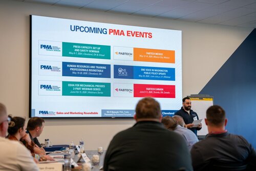 The image depicts a professional meeting or seminar setting. At the forefront, a man stands near a flip chart, possibly presenting or leading a discussion. He is holding a paper, suggesting he is referring to notes or an agenda. Around him, several people are seated at tables, engaged and listening, indicating a formal or business atmosphere.

The background features a large screen displaying a slide titled "UPCOMING PMA EVENTS." The slide lists several events organized by PMA (Precision Metalforming Association), each with specific details:

1. **Press Capacity: Set Up and Safety Seminar**
   - Date: May 9-10, 2023
   - Location: Cleveland, OH & Virtual
   - Partner: FABTECH

2. **Human Resources and Training Professionals Roundtable**
   - Date: May 25, 2023
   - Location: Cleveland, OH
   - Partner: One Voice

3. **OSHA for Mechanical Presses 2-Part Webinar Series**
   - Dates: June 7 and June 14, 2023
   - Partner: FABTECH

4. **One Voice Washington: Public Policy Update**
   - Date: June 8, 2023
   - Presenter: Omar Nashashibi, The Franklin Partnership

5. **FABTECH Canada**
   - Date: June 14-16, 2023
   - Location: Toronto, ON, Canada

The slide includes logos for PMA, FABTECH, and One Voice, indicating collaboration between these organizations. The room is well-lit, with a neutral color palette dominated by blues and whites, contributing to a professional and focused atmosphere.

The setting and content suggest this is a business or industry-related event, possibly a planning or informational session for upcoming conferences and seminars. The presence of industry-specific terminology and partner organizations indicates a specialized audience, likely professionals in the metalforming or manufacturing sectors.
