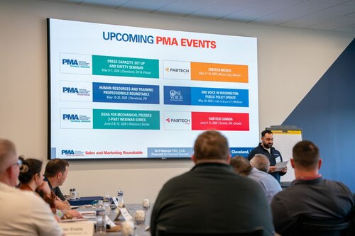 The image depicts a professional setting, likely a conference or seminar room, where a group of individuals is gathered for a presentation. The focal point is a large digital screen displaying a schedule titled "UPCOMING PMA EVENTS." The screen lists several events, including:

1. "PRESS CAPACITY, SET UP AND SAFETY SEMINAR" in Cleveland, OH.
2. "HUMAN RESOURCES AND TRAINING PROFESSIONALS ROUND TABLE" in Cleveland, OH.
3. "OSHA FOR MECHANICAL PRESSES 2 PART WEBINAR SERIES" online.
4. "FABTECH MEXICO" in Mexico City, Mexico.
5. "ONE VOICE WASHINGTON PUBLIC POLICY OUTLOOK" in Washington, DC.
6. "FABTECH CANADA" in Toronto, ON, Canada.

Logos for PMA (Precision Metalforming Association), FABTECH, and ONE VOICE are visible, indicating a focus on manufacturing and industry-related topics.

In the foreground, a speaker stands beside a flip chart, engaging with the audience, who are seated at tables. The audience appears attentive, suggesting an educational or informative atmosphere. The room is well-lit with natural and artificial lighting, contributing to a professional and focused environment.

The predominant colors are blue and white, creating a clean and corporate aesthetic. The setting and content suggest this is a business or industry-related event, likely aimed at professionals in the manufacturing sector. The overall atmosphere is formal and informative, emphasizing networking and knowledge sharing within the industry.