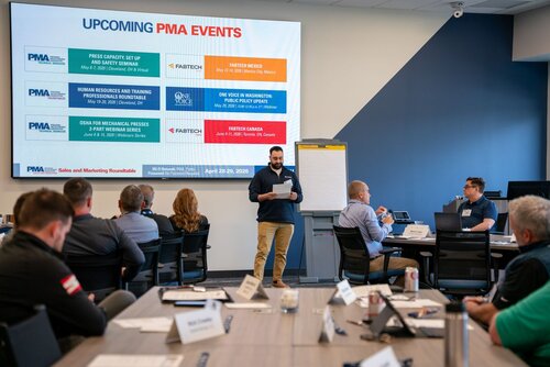 The image depicts a professional meeting or conference setting, likely a sales and marketing roundtable, as indicated by the text on the screen. The room is set up with a large presentation screen displaying upcoming PMA (Precision Metalforming Association) events. The screen lists various events with dates and locations, such as "Press Capacity Set Up and 3D Printing Seminar" and "Human Resources and Training Professionals Roundtable," among others. 

A man stands at the front of the room, holding a piece of paper, likely presenting or leading the discussion. Attendees are seated around a conference table, engaging with the presentation. Each participant has a nameplate, suggesting a formal or organized setting. The attendees appear attentive, with some taking notes or looking at the presenter.

The room is well-lit with natural and artificial lighting, creating a professional and focused atmosphere. The predominant colors are neutral, with shades of blue and gray on the walls and furniture, contributing to a corporate environment.

The setting suggests a business or industry-related event, with a focus on networking, education, and collaboration within the metalforming industry. The presence of specific event titles and the structured arrangement of participants highlight the organized nature of the gathering.