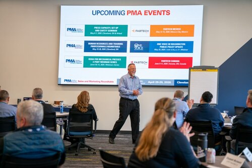 The image depicts a professional seminar or workshop setting. At the center, a speaker stands in front of a large screen displaying the title "UPCOMING PMA EVENTS" in bold, with "PMA" highlighted in red. The screen lists several events, including:

1. "PRESS CAPACITY, SETUP AND SAFETY SEMINAR" scheduled for May 24-25, 2023, in Cleveland, OH.
2. "HUMAN RESOURCES AND TRAINING PROFESSIONALS ROUNDABLE" on May 31, 2023, via webinar.
3. "OSHA FOR MECHANICAL PRESSES" as part of a 2023 webinar series.
4. "FABTECH MEXICO" on May 16-18, 2023, in Mexico City.
5. "ONE VOICE IN WASHINGTON POLICY UPDATE" on June 6, 2023, via webinar.
6. "FABTECH CANADA" on June 14-16, 2023, in Toronto.

The bottom of the screen shows the date range "April 28-29, 2023."

The speaker is casually dressed and appears to be engaging with the audience, who are seated at tables. Attendees are seen taking notes and some are clapping, indicating an interactive and positive atmosphere. The room is well-lit, with a modern and professional ambiance. The presence of a flip chart suggests a focus on collaborative learning or discussion.

The event seems to be related to the manufacturing or industrial sector, given the context of the listed events and the mention of "FABTECH," a well-known trade show for metal forming, fabricating, welding, and finishing.

Overall, the image conveys a theme of professional development and industry networking, with a focus on upcoming events and opportunities for learning and collaboration within the manufacturing industry.