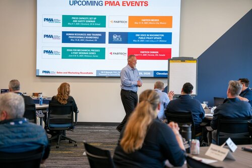 The image depicts a professional setting, likely a conference or seminar, with a focus on upcoming events organized by PMA (Precision Metalforming Association). A man is standing and speaking in front of an audience, suggesting he is leading a presentation or discussion. The audience is seated at tables, indicating a formal meeting environment.

A large screen at the front displays information about "UPCOMING PMA EVENTS," listing several events with their dates and locations. These include:

1. Press Capacity, Set Up, and Safety Seminar in Cleveland, OH.
2. Human Resources and Training Professionals Roundtable in Cleveland, OH.
3. OSHA for Mechanical Presses 2-Part Webinar Series.
4. FABTECH Mexico in Mexico City, Mexico.
5. One Voice in Washington: Public Policy Update in Washington, D.C.
6. FABTECH Canada in Toronto, ON, Canada.

The screen is a key focal point, providing detailed event information and logos for PMA and FABTECH, suggesting a connection to the metalforming industry.

The setting is a modern conference room with a professional atmosphere. The lighting is bright and conducive to presentations. Attendees are casually dressed, reflecting a business-casual dress code typical of industry events.

The presence of a flip chart next to the speaker suggests interactive elements or note-taking during the session. The overall mood is focused and educational, with participants engaged in learning about industry events and updates.

This image captures a typical industry conference scene, highlighting the importance of networking and professional development within the metalforming sector.