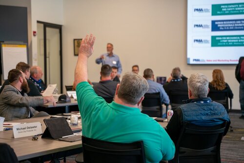 The image depicts a professional meeting or conference setting in a modern office environment. Several individuals are seated around a conference table, engaged in a discussion. One person in the foreground, wearing a teal shirt, is raising their hand, possibly to ask a question or contribute to the conversation. 

The room is well-lit, with natural and artificial lighting, creating a bright and focused atmosphere. A large screen at the front displays a presentation with text and logos, suggesting an organized agenda. The screen content includes dates and titles, indicating upcoming events or sessions. 

Participants are dressed in business casual attire, and there are nameplates on the table, one of which reads "Michael Dilawyer." The presence of laptops, notebooks, and beverages like a can of soda suggests a collaborative and informal yet professional setting.

The atmosphere is one of active engagement and learning, typical of a workshop or seminar. The room's decor is minimalistic, with neutral colors, enhancing the focus on the meeting's content. This image captures a moment of interaction and exchange of ideas, characteristic of corporate or educational gatherings.