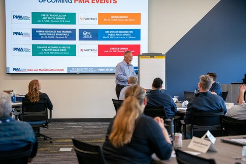 The image depicts a professional meeting or seminar taking place in a modern conference room. A group of individuals is seated around tables, attentively facing a presenter. The presenter, a man in a light blue shirt, stands beside a flip chart, actively engaging with the audience. 

On the wall behind him is a large screen displaying a presentation slide titled "UPCOMING PMA EVENTS." The slide lists several events with dates and locations, including "PRESS CAPACITY, SET UP AND EFFICIENCY SEMINAR," "HUMAN RESOURCES AND TRAINING PROFESSIONALS NETWORKING," "OSHA PSM MECHANICAL PRESS & PART METAL STAMPING SEMINAR," "FABTECH MEXICO," "ONE VOICE WASHINGTON PUBLIC POLICY UPDATE," and "FABTECH CANADA." The slide features logos of PMA and FABTECH, indicating a connection to these organizations.

The room is well-lit with a combination of natural and artificial lighting, creating a professional and focused atmosphere. Attendees are seated at round tables, each with name tags and notepads, suggesting an organized and formal setting. The predominant colors in the room are neutral tones, with accents of blue and red from the presentation slide.

The setting and content suggest a business or industry-related event, likely focusing on manufacturing or engineering topics, given the mention of mechanical presses and metal stamping. The atmosphere is one of learning and collaboration, with participants likely engaging in discussions related to the topics presented.