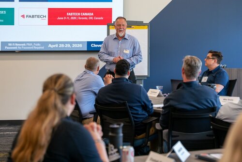 The image depicts a professional meeting or conference setting, likely a seminar or workshop. A group of individuals is seated around tables, engaging in a presentation. The presenter, a middle-aged man, stands at the front, speaking to the audience. He is dressed in a light blue shirt and dark pants, exuding a professional demeanor. Behind him, a screen displays slides with text related to events, including "FABTECH CANADA" scheduled for June 11-13, 2024, in Toronto, ON, Canada, and a future event in Cleveland, OH, on April 28-29, 2026. 

The room has a modern and professional ambiance, with a mix of blue and neutral tones. A flip chart is visible next to the presenter, suggesting a focus on interactive or educational content. Attendees, mostly dressed in business casual attire, are attentively listening, some taking notes. The setting conveys a sense of collaboration and learning, typical of industry conferences or corporate training sessions. The atmosphere is focused and engaged, indicative of a professional development or networking event.