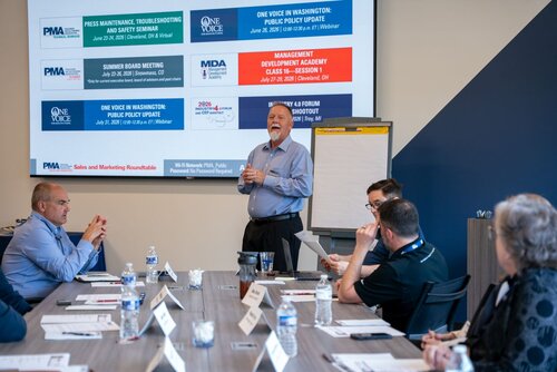 The image depicts a professional meeting or seminar taking place in a conference room. At the center, a man is standing and speaking to a group of seated individuals, suggesting he is leading the discussion or presentation. The participants are seated around a table, attentively listening, with some taking notes. The table is equipped with water bottles, papers, and pens, indicating a formal setting.

In the background, a large screen displays several event titles and dates, such as "Press Maintenance, Troubleshooting and Safety Seminar," "Summer Board Meeting," and "Management Development Academy Class 20 - Session 1." These suggest the context is related to professional development or industry-specific training, possibly in manufacturing or public policy.

The setting is well-lit, with a modern and professional atmosphere. The walls are painted in a combination of white and dark blue, adding to the corporate environment. A flip chart is also visible, hinting at interactive elements in the presentation.

The individuals are dressed in business casual attire, reinforcing the professional tone. The overall mood is focused and engaged, indicative of a collaborative and informative session. The presence of logos such as "PMA" and "One Voice" suggests affiliations with specific organizations or initiatives, likely related to manufacturing or policy advocacy.