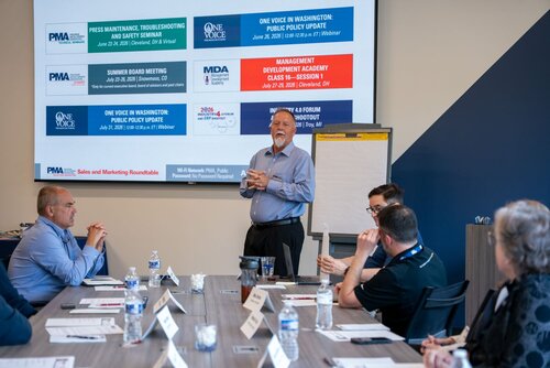 The image depicts a professional meeting or seminar taking place in a conference room. A group of individuals is seated around a table, which is covered with documents, notepads, and water bottles, indicating a formal setting. The attendees are dressed in business attire, suggesting a corporate or industry-related event.

A man stands at the front of the room, speaking to the group. He appears to be leading the session, possibly as a presenter or facilitator. Behind him is a large display board with several banners and logos. The banners contain text related to different events and organizations, such as "PMA," "One Voice," and "MDA." These likely represent industry associations or initiatives.

The banners list various events and updates, including "Press Maintenance Troubleshooting," "Summer Board Meeting," and "Management Development Academy Class." This suggests the meeting is focused on industry-specific topics, possibly related to manufacturing or business management.

The room is well-lit, with a modern and professional atmosphere. The presence of a flip chart next to the speaker further emphasizes the educational or training aspect of the event. The overall mood is one of focus and engagement, typical of a professional development or strategic planning session.