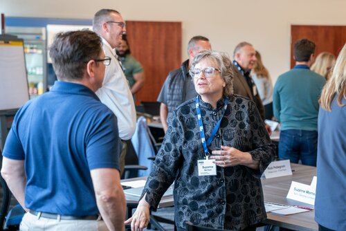 The image depicts a professional networking or workshop event in an indoor setting, likely a conference room. The atmosphere is lively and engaged, with several individuals interacting and conversing. The attendees are dressed in business casual attire, suggesting a formal yet relaxed environment.

A woman in the foreground, wearing glasses and a patterned dark blouse, is speaking with a man in a navy blue polo shirt. She has a name badge on a lanyard, indicating her participation in the event. Other attendees are visible in the background, engaged in similar conversations, creating a sense of community and collaboration.

The room is well-lit with natural and artificial lighting, enhancing the clarity of the scene. Tables are arranged with name tags, indicating a structured setup for discussions or presentations. The presence of a flip chart or whiteboard suggests the possibility of presentations or brainstorming sessions.

Overall, the image conveys a professional and collaborative atmosphere, typical of networking events, workshops, or seminars aimed at fostering connections and sharing knowledge.