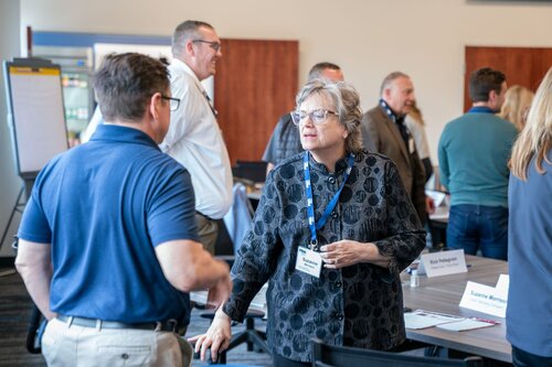 The image depicts a professional networking or business meeting setting. In the foreground, two individuals are engaged in conversation. The woman is wearing a patterned black blouse and glasses, with a name badge and lanyard around her neck, suggesting a formal event. The man facing her is dressed in a blue polo shirt and beige pants, also wearing a name badge.

In the background, several other people are interacting, indicating a lively and social atmosphere. A flip chart is visible on the left side, hinting at a presentation or brainstorming session. Tables are arranged with name cards, water bottles, and papers, reinforcing the formal and organized nature of the event.

The room is well-lit with natural and artificial lighting, creating a bright and welcoming environment. The overall atmosphere is professional and collaborative, typical of a conference, seminar, or workshop. The attire of the participants is business casual, aligning with a corporate or educational setting.