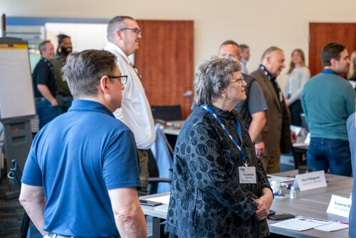 The image depicts a professional setting, likely a conference or workshop, with several individuals standing and engaging in conversation. The participants are dressed in business casual attire, suggesting a formal yet relaxed atmosphere. Prominent in the foreground is a woman wearing a patterned black jacket and glasses, looking attentively towards the front of the room. Beside her, a man in a blue polo shirt is also focused in the same direction. Both individuals wear name tags, indicating an organized event where networking or identification is necessary.

In the background, more attendees are visible, some standing and others seated at tables. The room is well-lit with natural light, enhancing the clarity and warmth of the setting. Tables are arranged with nameplates and materials, typical of a seminar or meeting environment. The presence of a flip chart and a projector screen suggests presentations or collaborative activities are part of the event.

The overall mood is professional and engaged, with participants appearing attentive and interested. The setting and arrangement of elements indicate a structured event aimed at learning, discussion, or professional development.