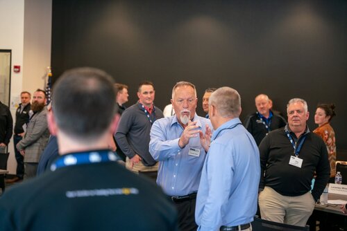 The image depicts a professional networking or business event taking place in a conference room. The setting is formal, with individuals dressed in business attire, including button-up shirts and slacks. The room has a modern design with a dark wall in the background, contributing to a professional atmosphere. 

In the foreground, two men are engaged in conversation. One man, with gray hair and a beard, is gesturing with his hands, suggesting an animated discussion or explanation. The other man is listening attentively. Both are wearing name tags, indicating a formal event where identification is necessary, such as a conference or seminar.

Several other individuals are visible in the background, also engaged in conversations, suggesting a networking session. An American flag is partially visible on the left side, hinting at a location in the United States or an event with a national context.

The lighting is bright and even, typical of indoor conference settings, which enhances the clarity of the scene. The overall atmosphere is professional and collaborative, indicative of a business or educational event focused on interaction and exchange of ideas.