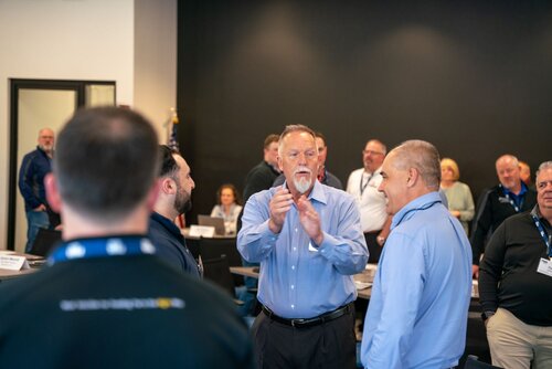 The image depicts a group of people gathered in what appears to be a professional or corporate setting, likely a conference or meeting room. The central figure, an older man with a beard, is engaged in animated conversation with two other men. He is gesturing with his hands, suggesting he is explaining or emphasizing a point. The individuals around him are dressed in business casual attire, with collared shirts being predominant.

In the background, several other people are visible, some seated and others standing, suggesting a break or networking session during a conference or workshop. The room is well-lit, with a neutral color palette dominated by blues and blacks, contributing to a professional atmosphere. The presence of name tags on lanyards indicates that this is an organized event, possibly involving multiple participants from different organizations.

An American flag is partially visible in the background, hinting at a formal or official setting. The overall mood is one of engagement and discussion, typical of a collaborative or educational environment. The setting and attire suggest a focus on business or professional development, with an emphasis on communication and interaction among participants.