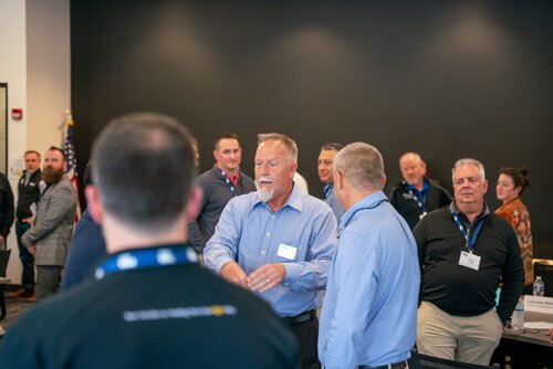 The image depicts a professional gathering or networking event, likely a conference or business meeting. The setting appears to be indoors, with a dark wall in the background that contrasts with the well-lit room. The lighting is bright and evenly distributed, suggesting a formal environment.

Several individuals, mostly men, are engaged in conversation. They are dressed in business attire, including suits and collared shirts, indicating a professional or corporate atmosphere. One man in the center, wearing a light blue shirt and a name tag, appears to be speaking or leading a discussion, suggesting he might be a key speaker or facilitator.

In the background, an American flag is visible, hinting at a U.S.-based event or organization. Name tags worn by attendees imply a formal networking or identification process typical of conferences. 

The mood of the image is professional and engaged, with participants appearing attentive and interactive. The setting suggests a focus on business, networking, or professional development.