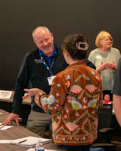 The image depicts a casual, indoor meeting or workshop setting. In the foreground, a man and a woman are engaged in conversation. The man, wearing a dark jacket with a name tag, appears to be smiling, suggesting a friendly interaction. The woman, seen from the back, is wearing a distinctive brown jacket adorned with colorful floral embroidery, adding a vibrant touch to the scene.

In the background, another woman stands, also smiling, contributing to the overall positive and welcoming atmosphere. She wears a light-colored top and has a name tag, indicating this is a professional or networking event. A table is visible with papers, a water bottle, and a Coca-Cola can, suggesting a casual and informal environment conducive to discussion and collaboration.

The setting is well-lit, with neutral-colored walls and furniture, creating a professional yet relaxed ambiance. The presence of name tags and documents indicates an organized event, possibly a seminar, workshop, or business meeting. The image conveys a theme of communication and engagement, with a focus on interpersonal interaction.