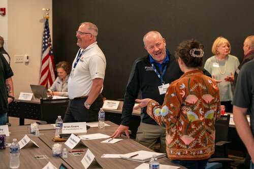 The image depicts a professional meeting or workshop setting. Several individuals are engaged in conversation around a conference table. The room has a formal atmosphere, indicated by the presence of name tags and a structured seating arrangement with nameplates. 

In the foreground, a man wearing a dark jacket with a name tag labeled "Brian Evans" is smiling and interacting with a woman in a colorful, patterned jacket. Their interaction appears friendly and animated, suggesting a positive and engaging atmosphere.

In the background, an American flag is visible, hinting at a U.S. location or organization. Several other individuals are also engaged in discussions, contributing to a collaborative and dynamic environment. The room is well-lit, with neutral tones and a focus on professional decorum.

The setting suggests a business or educational context, possibly a seminar, training session, or networking event. Bottles of water and papers are placed on the table, indicating an organized and prepared meeting space. The overall mood is professional yet approachable, fostering interaction and exchange of ideas.