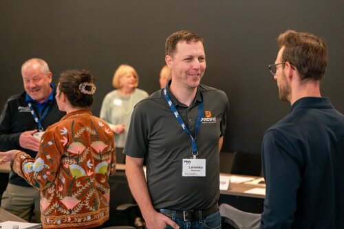 The image depicts a professional networking or meeting event, likely in a conference room setting. Several individuals are engaged in conversation, suggesting a collaborative or social atmosphere. 

In the foreground, two men are talking. One wears a gray polo shirt with a logo and a name badge that reads "Laramay," indicating a formal or organized event. The other man is dressed in a dark shirt and glasses, suggesting a casual yet professional dress code.

In the background, a woman with a floral-patterned jacket and a man in a black jacket are also conversing, adding to the dynamic interaction among attendees. The room has a neutral color scheme with dark walls, which enhances the focus on the participants.

The lighting is bright and even, creating a welcoming and open environment. The overall atmosphere is one of professionalism and engagement, typical of business meetings or networking events. The presence of name badges and logos suggests a corporate or industry-specific gathering, possibly related to a company or organization named "PRIME."

The image captures the essence of professional networking, highlighting interpersonal communication and the exchange of ideas in a formal setting.