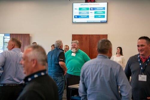 The image depicts a professional networking or business event taking place in a conference room. Several individuals are engaged in conversation, suggesting a casual and interactive atmosphere. The participants are dressed in business casual attire, with some wearing name tags, indicating a formal gathering or seminar.

In the background, a digital screen displays a slide titled "UPCOMING PMA EVENTS," listing various events with associated dates and details. This suggests the event is related to the PMA (Precision Metalforming Association) or a similar organization, focusing on industry-specific topics.

The room is well-lit with neutral-colored walls, contributing to a professional and welcoming environment. The presence of tables and chairs arranged for group discussions indicates a setting designed for collaboration and networking.

Overall, the image conveys a theme of professional development and industry engagement, with an emphasis on community building and information sharing within a specific professional field.