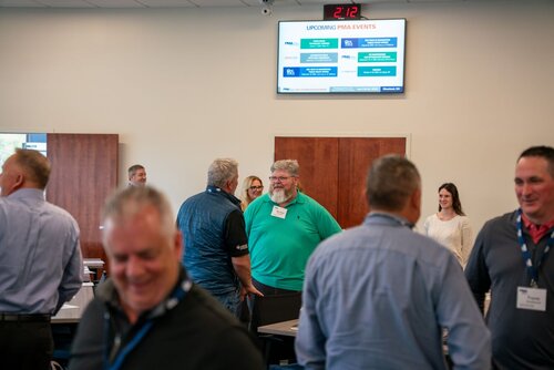 The image depicts a professional networking or business event taking place in a conference room. Several individuals are engaged in conversation, suggesting a social or networking atmosphere. The attendees are dressed in business casual attire, with name tags visible, indicating a formal gathering or seminar.

In the background, a digital screen displays a schedule titled "UPCOMING PMA EVENTS," listing various events and activities, which suggests the setting is related to a professional organization or association. The screen also shows a digital clock reading "2:12," indicating the time of day.

The room has a neutral color palette with beige walls and wooden doors, contributing to a professional and organized environment. The lighting is bright and even, typical of indoor conference settings.

The mood is lively and engaging, with participants smiling and interacting, reflecting a positive and collaborative atmosphere. The event appears to be focused on networking, professional development, or industry-specific discussions, as indicated by the formal setting and the presence of informational displays.