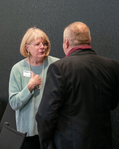 The image depicts two individuals engaged in conversation in what appears to be a professional or formal setting. The woman on the left is wearing a light blue sweater and a pearl necklace, with glasses and short blonde hair. She has a name tag on her sweater, indicating a formal or networking event. The man on the right is wearing a black suit with a maroon shirt, and his back is partially turned towards the camera, making his face less visible.

The background features a dark, textured wall, contributing to a neutral and understated atmosphere. The lighting is soft and even, suggesting an indoor environment, possibly a conference or meeting room. The interaction between the two individuals seems earnest and engaged, with the woman gesturing with her hand on her chest, indicating a personal or meaningful exchange.

The setting and attire suggest a professional gathering, such as a business conference, seminar, or networking event. The overall mood is formal yet approachable, emphasizing professional interaction and communication.